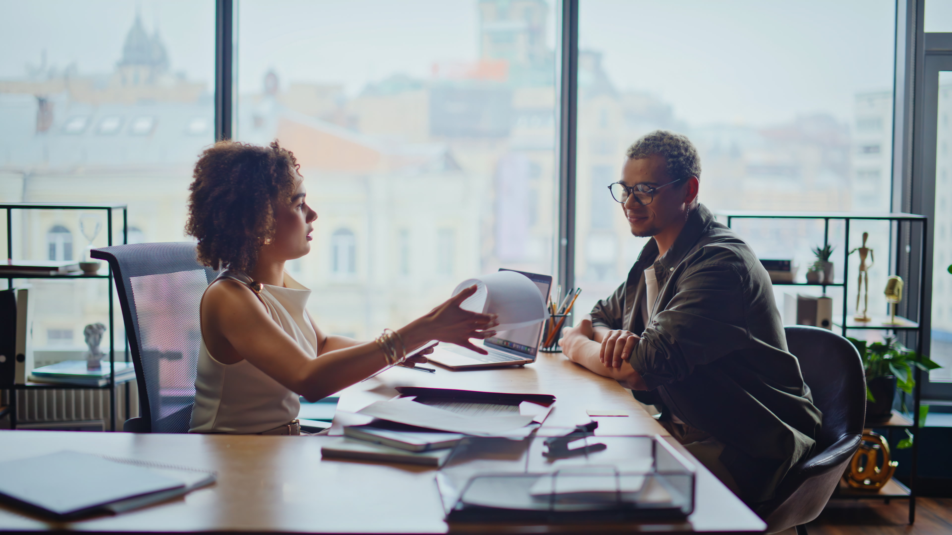 Two young colleagues discussing paperwork office desk, woman handing documents to man in glasses. Multiethnic business partners reviewing contract, focused negotiation meeting corporate teamwork.